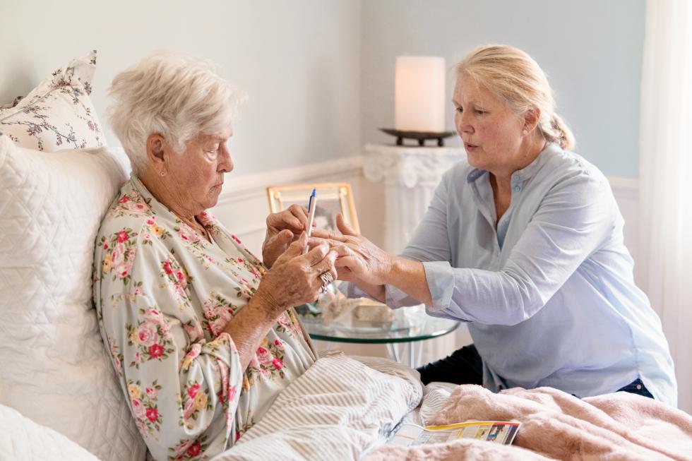 Frau reicht einer älteren Dame im Bett einen Kugelschreiber